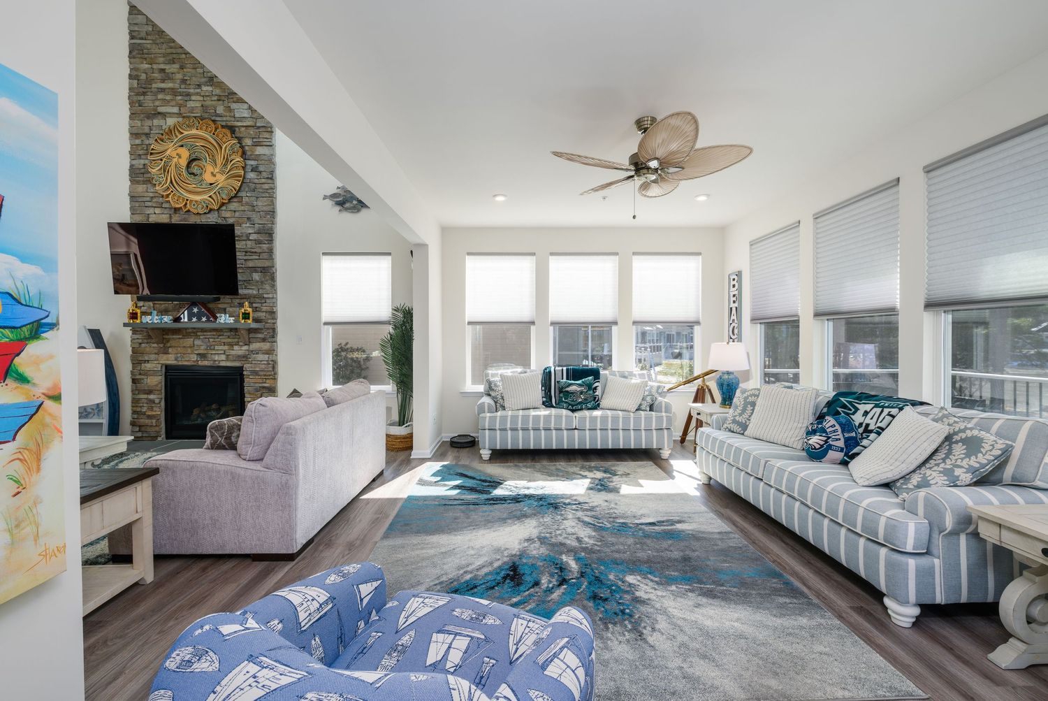 Bright coastal living room with stone fireplace, ceiling fan, blue area rug, and large windows flooding the space with natural light.