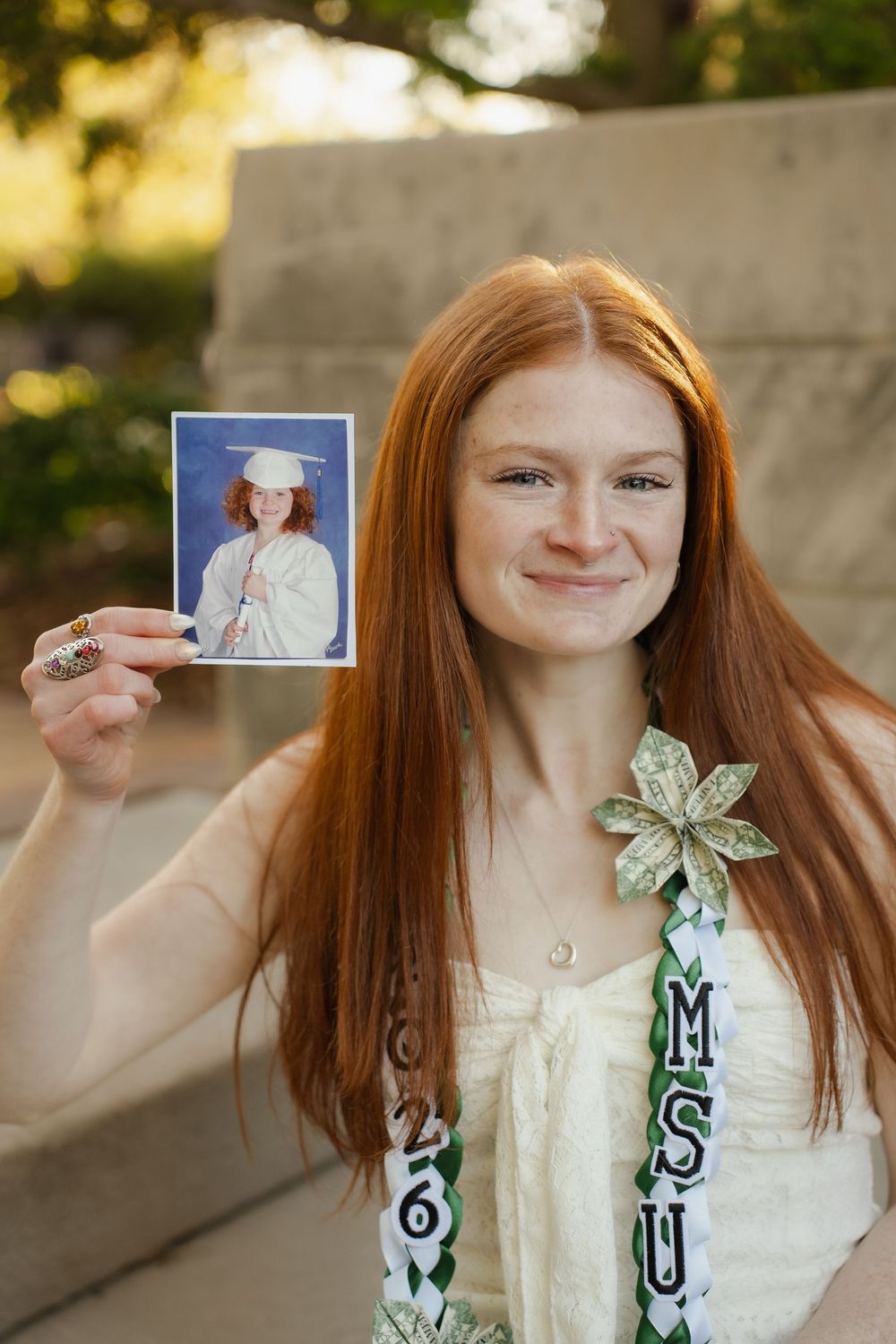 Red-haired graduate holding childhood graduation photo, wearing MSU sash and corsage, smiling outdoors.