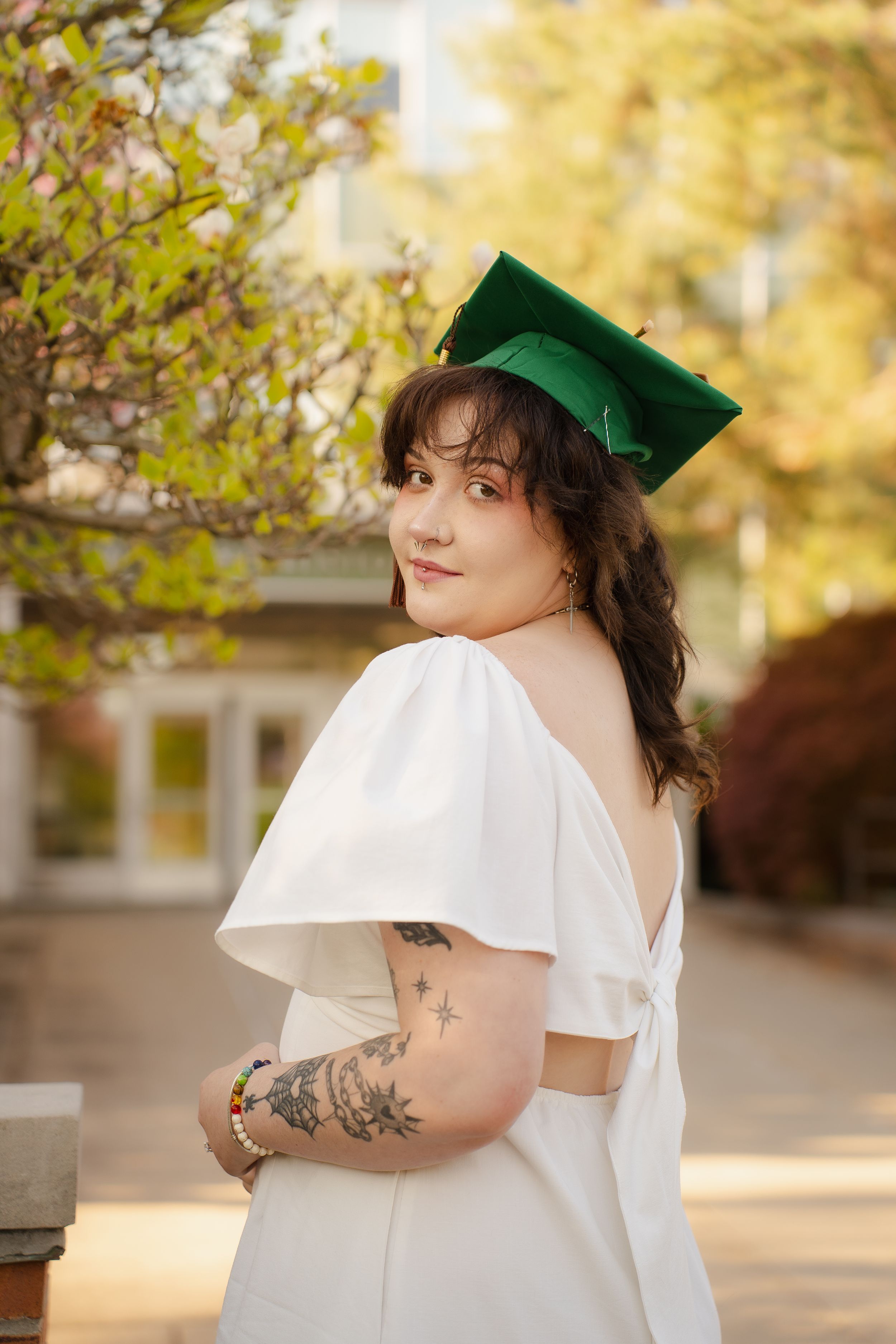 Young woman in white backless dress and green graduation cap poses outdoors, showcasing arm tattoos, celebrating her graduation.