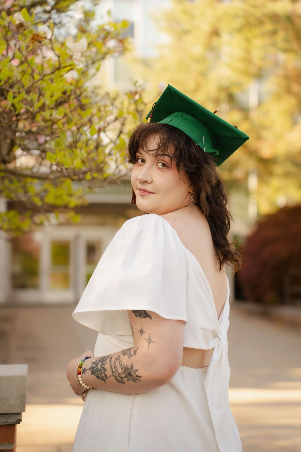 Young woman in white backless dress and green graduation cap poses outdoors, showcasing arm tattoos, celebrating her graduation.