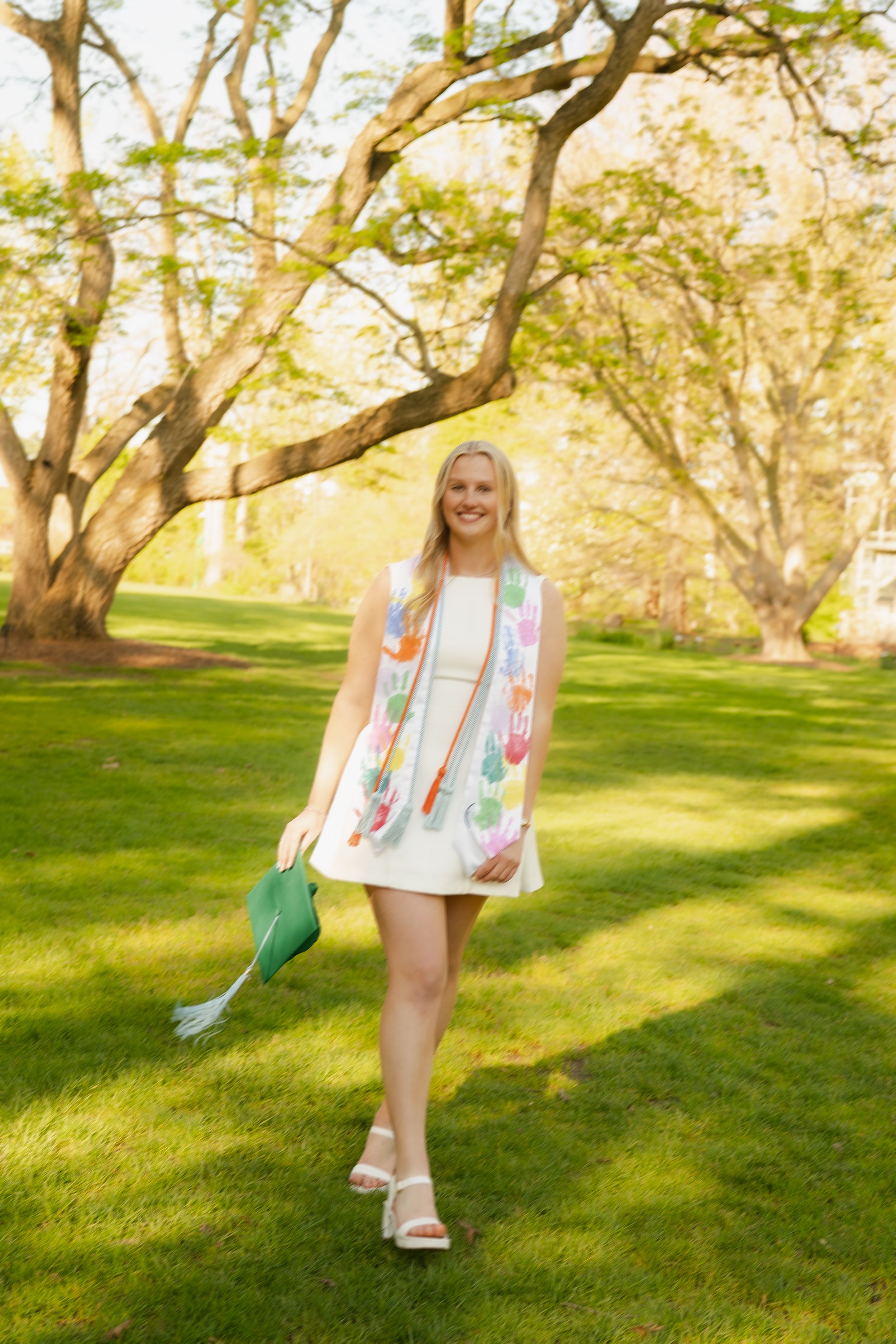Smiling graduate in white dress holding green cap walks through sunny park with colorful stole.
