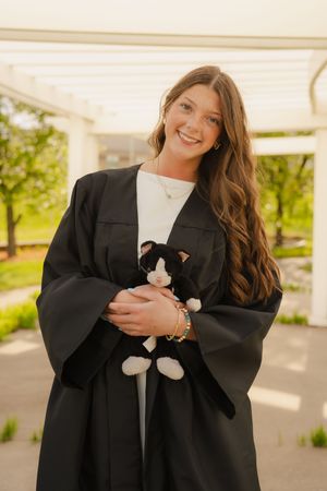 Smiling graduate in black gown holds a tuxedo cat stuffed animal outdoors near a pergola.