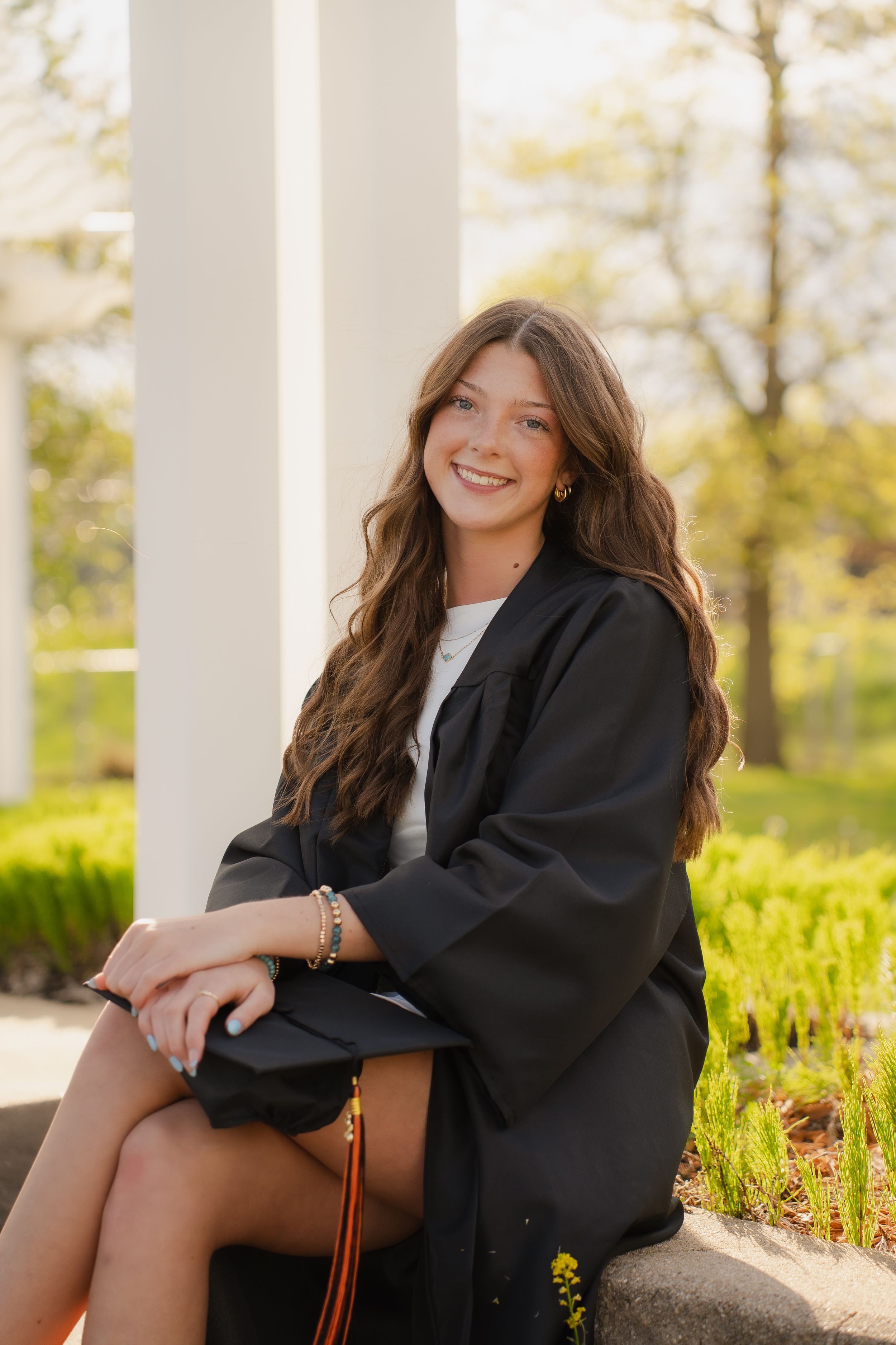Smiling young woman in black graduation gown sitting outdoors near white columns with green foliage background.