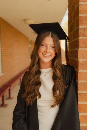 Smiling young woman in black graduation cap and gown leaning against brick wall outdoors.