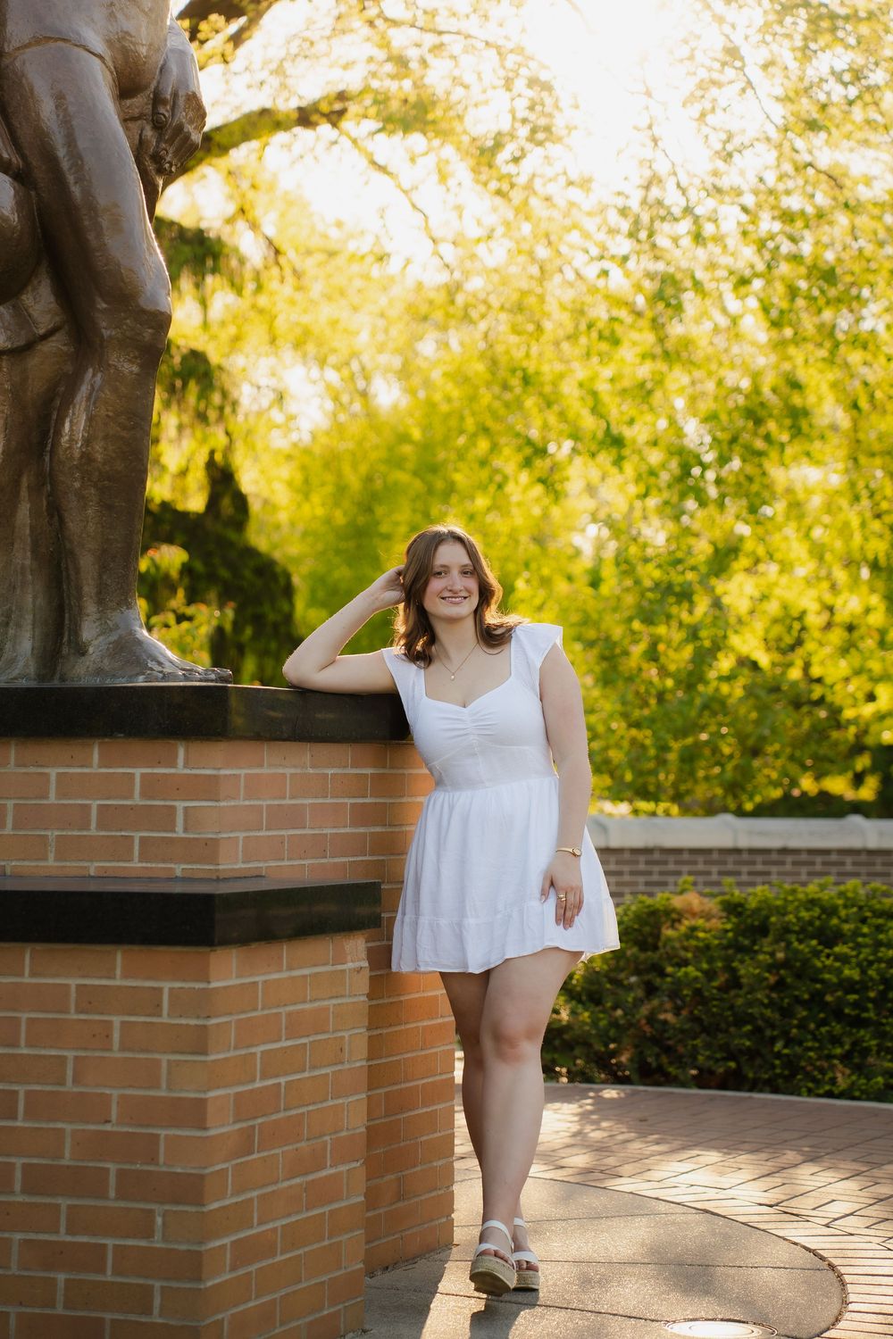 Young woman in white dress posing by brick statue base in sunny garden setting.