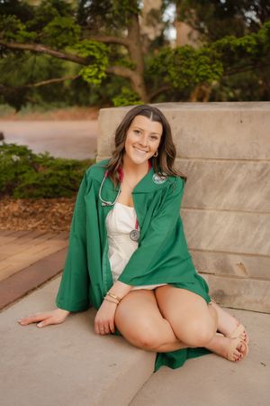 Smiling young woman in green graduation gown with stethoscope sits outdoors on stone steps among lush trees.