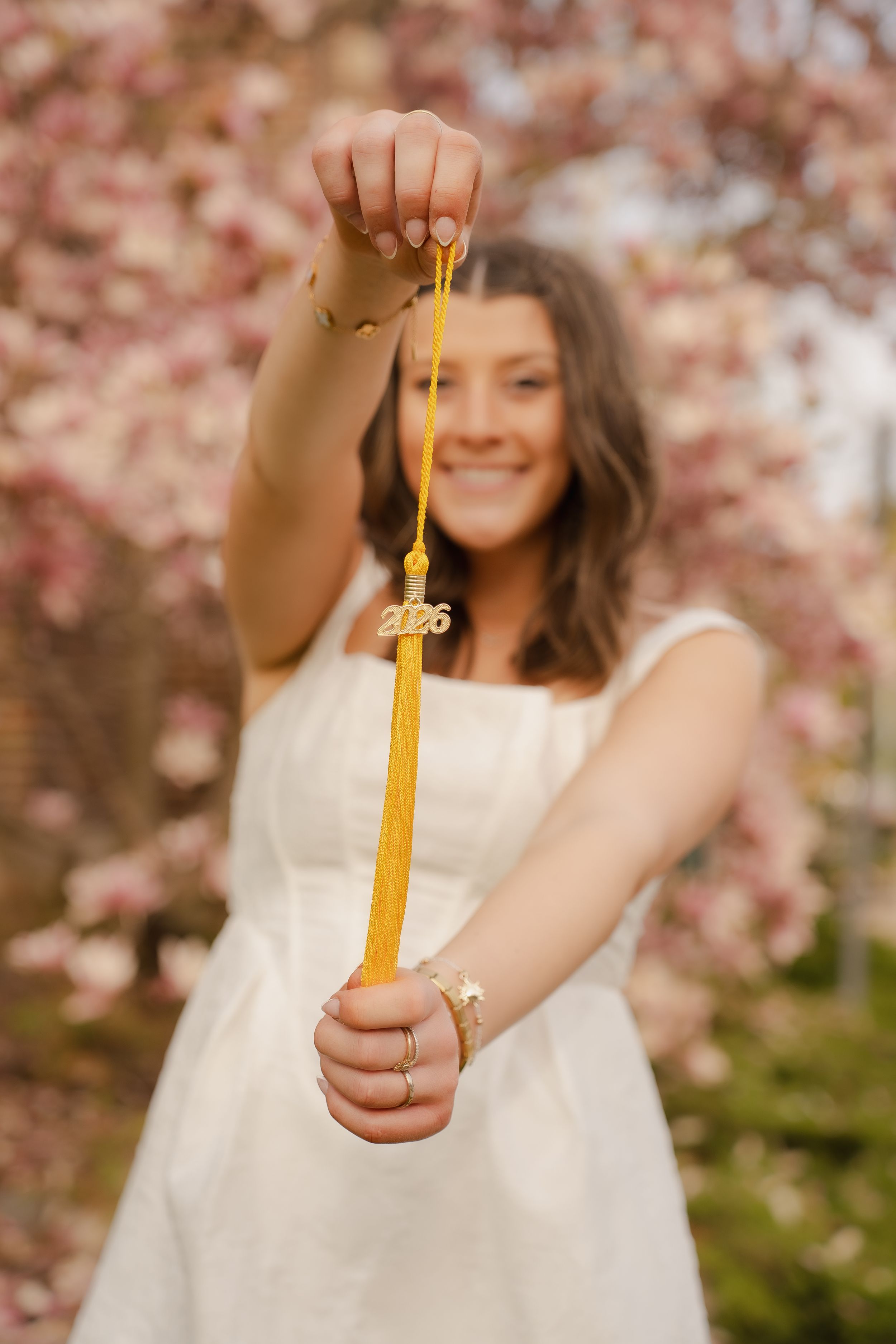 Smiling graduate in white dress holds gold 2026 tassel toward camera with blooming pink cherry trees in background.