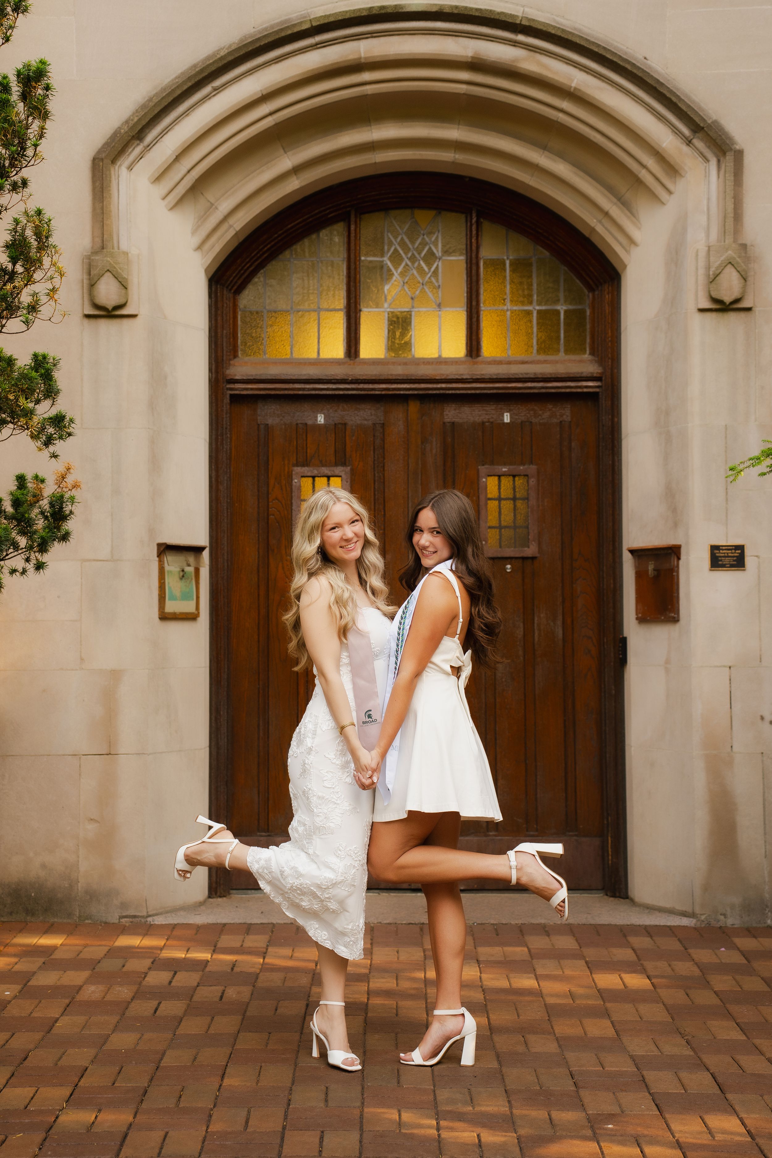 Two young women in white dresses and heels pose back-to-back holding hands before an ornate wooden arched doorway.