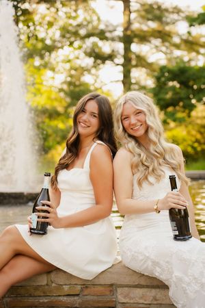 Two smiling women in white dresses sitting by a fountain, holding bottles, with lush green trees in the background.