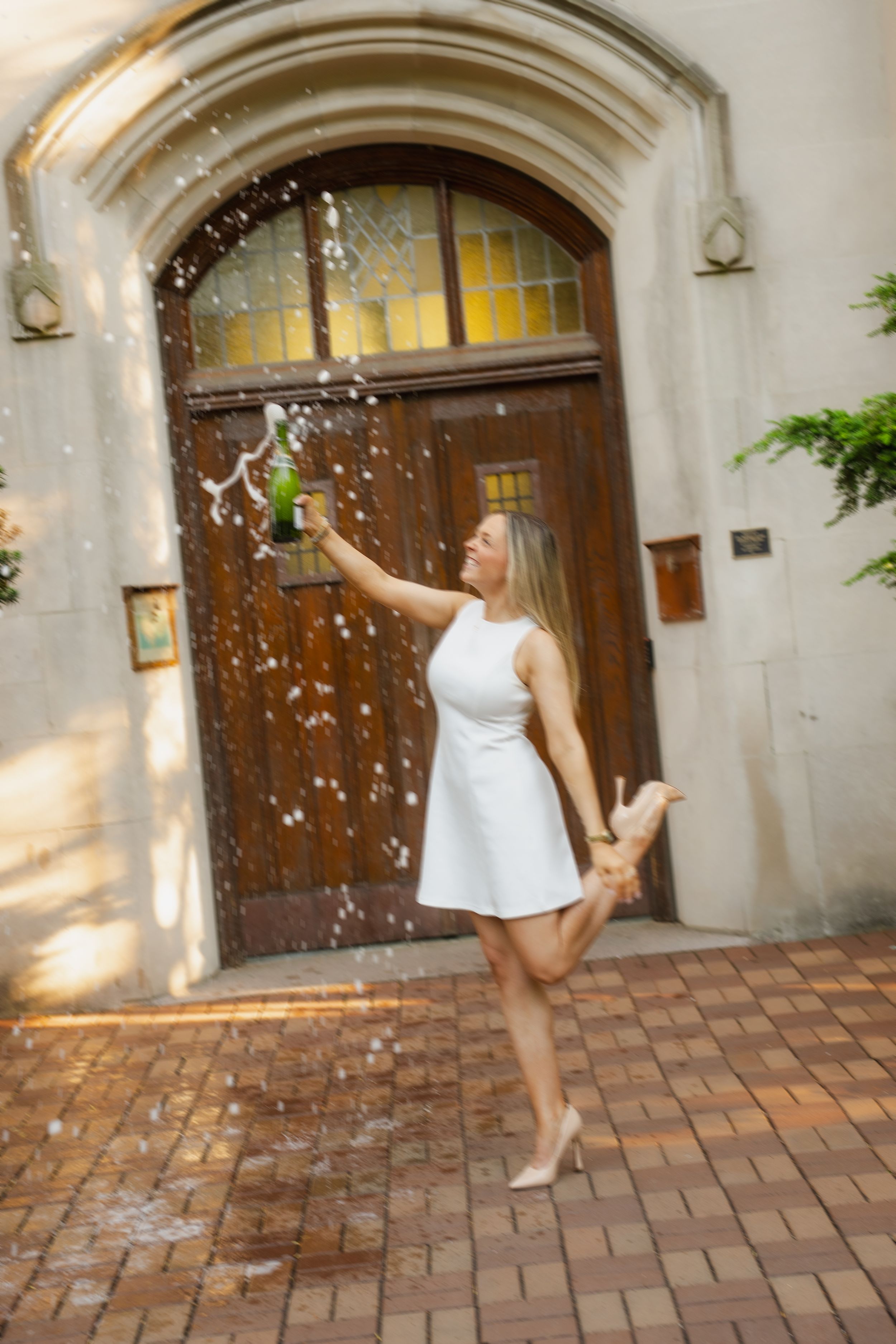 Woman in white dress celebrating with champagne bottle outdoors near ornate wooden door on brick patio.
