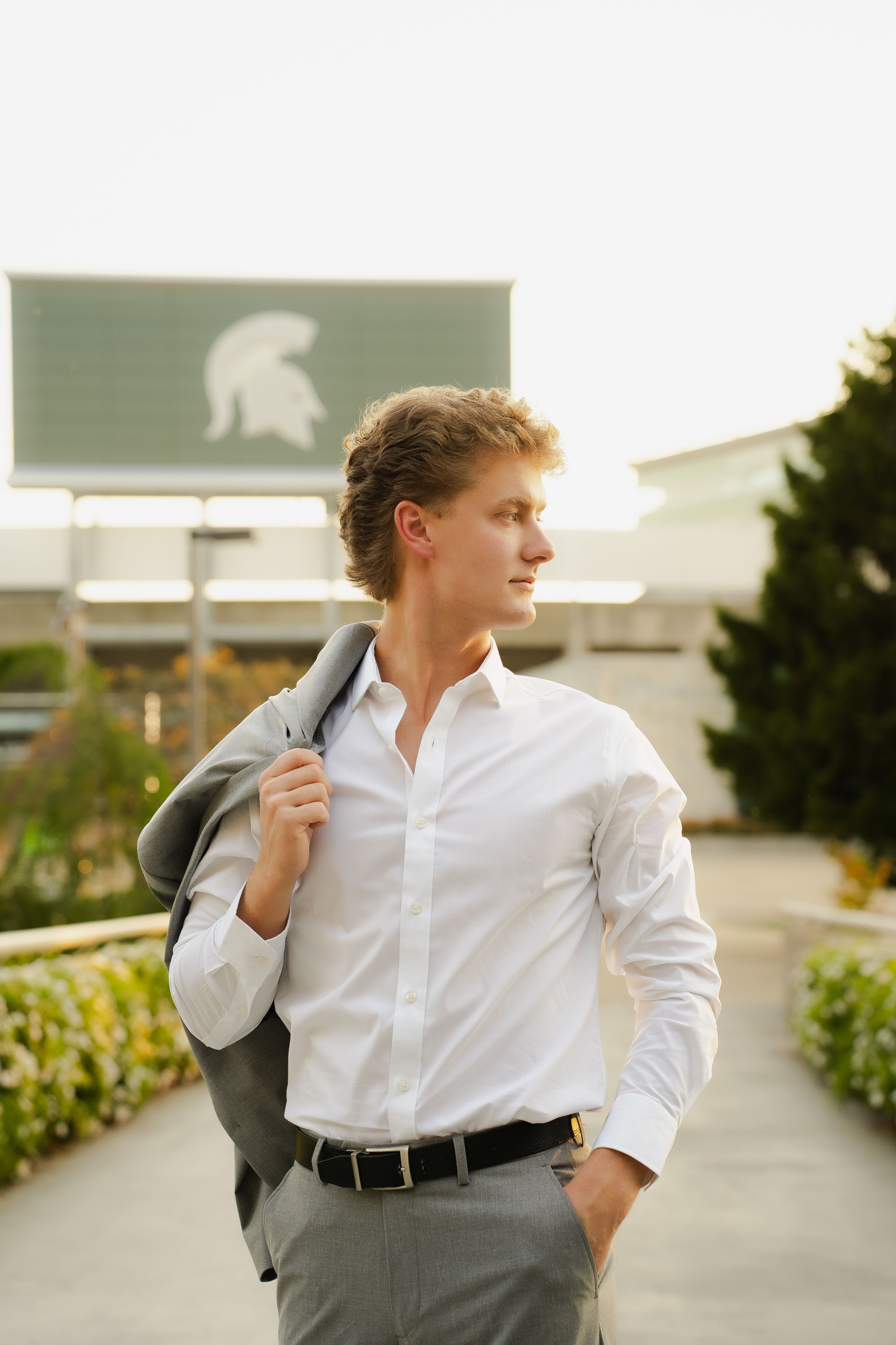 Young man in a white dress shirt and gray pants holds a blazer over his shoulder near a stadium sign with a Spartan helmet logo.
