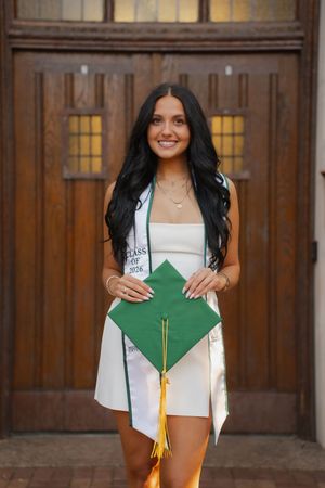 Smiling graduate in white dress holding green cap with gold tassel and Class of 2025 sash before wooden doors.