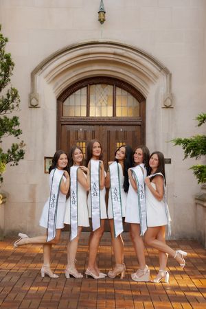 Six young women in white dresses and Greek sorority sashes pose together in front of an ornate stone building entrance.