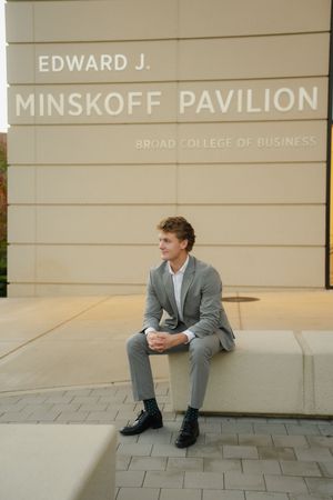 Young man in a gray suit sits on a stone bench outside the Edward J. Minskoff Pavilion at the Broad College of Business.