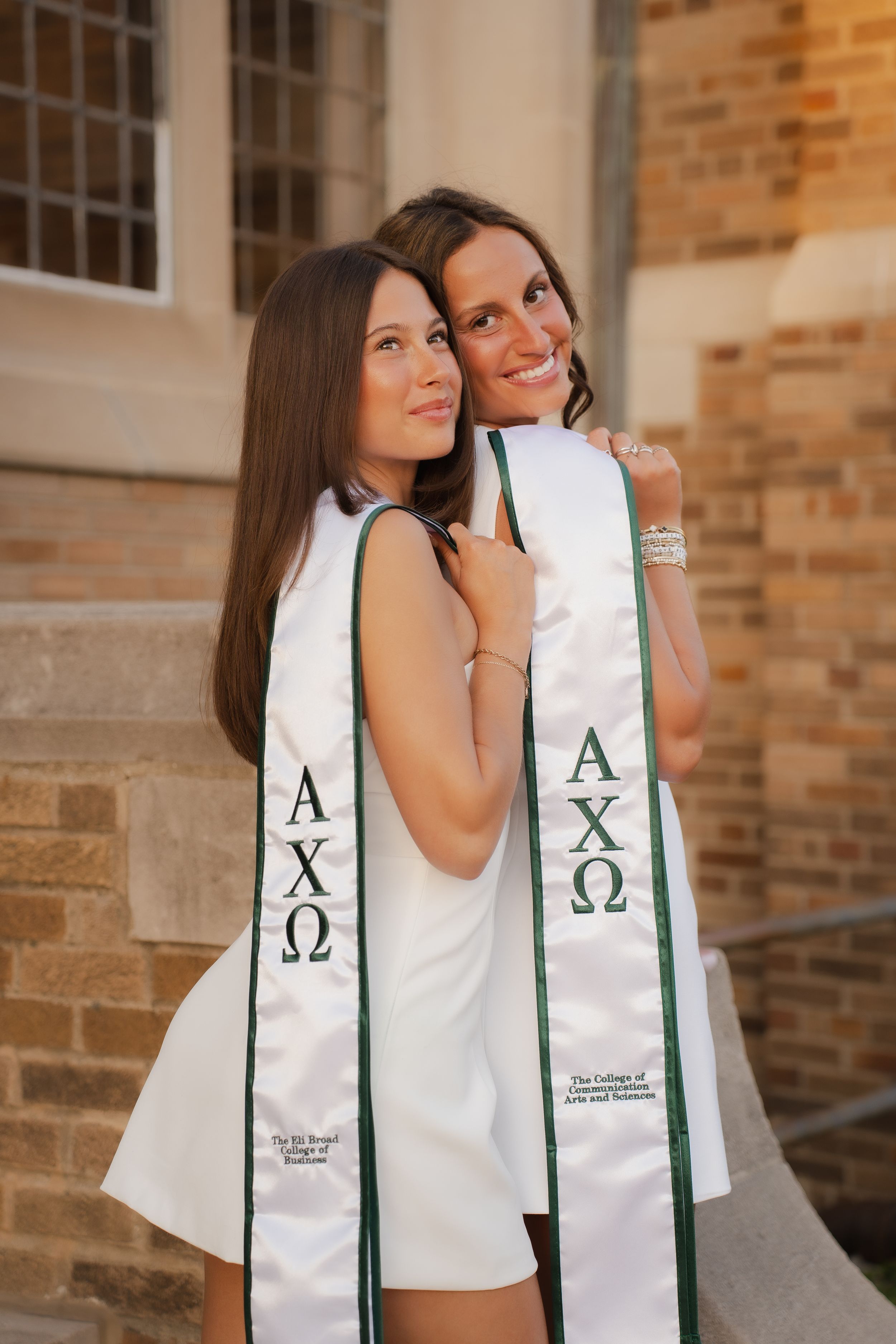 Two women wearing Alpha Chi Omega graduation stoles pose together outdoors near a brick building.
