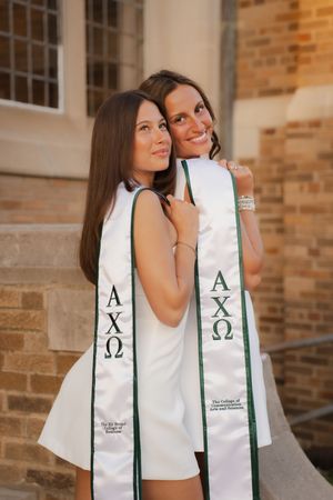 Two women wearing Alpha Chi Omega graduation stoles pose together outdoors near a brick building.