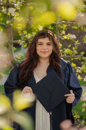 Young woman in navy graduation gown holding cap outdoors among blooming spring trees, smiling proudly.