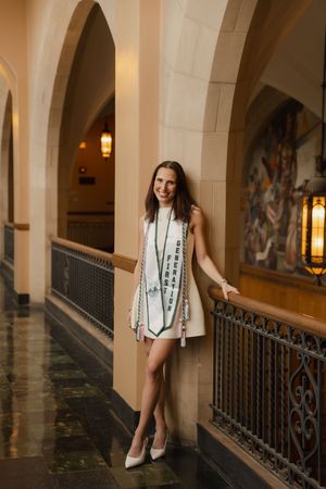 Smiling graduate in white dress and green graduation stole poses in elegant arched hallway with ornate decor.