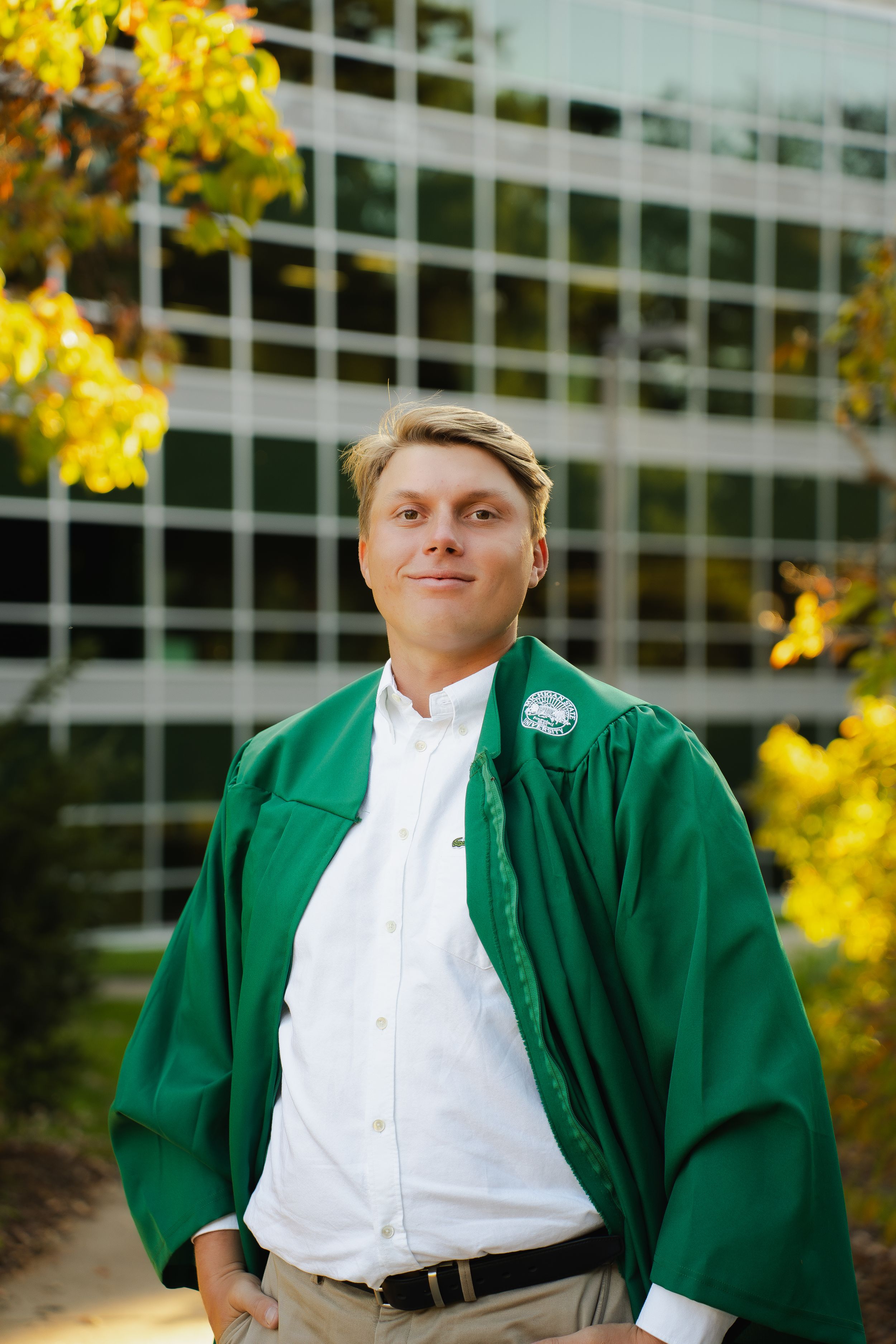 Smiling graduate in a green gown over a white shirt and khakis poses outdoors near a glass building and golden autumn leaves.