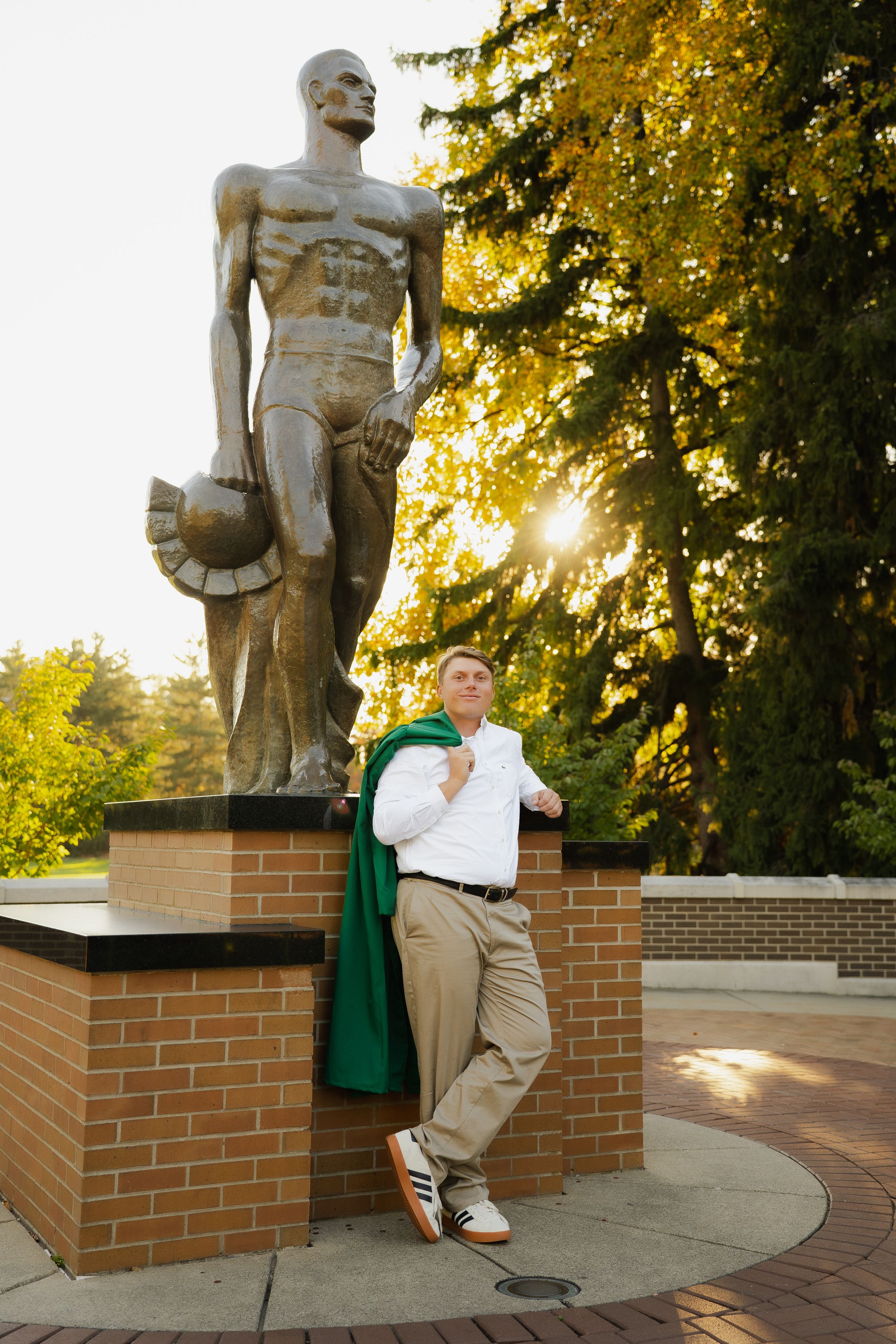 Man in a white shirt and khakis with a green jacket draped over his shoulders leans on a brick base beside a bronze statue at sunset.