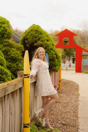 Smiling blonde woman in white lace dress posing by colorful crayon fence posts with red schoolhouse arch backdrop.
