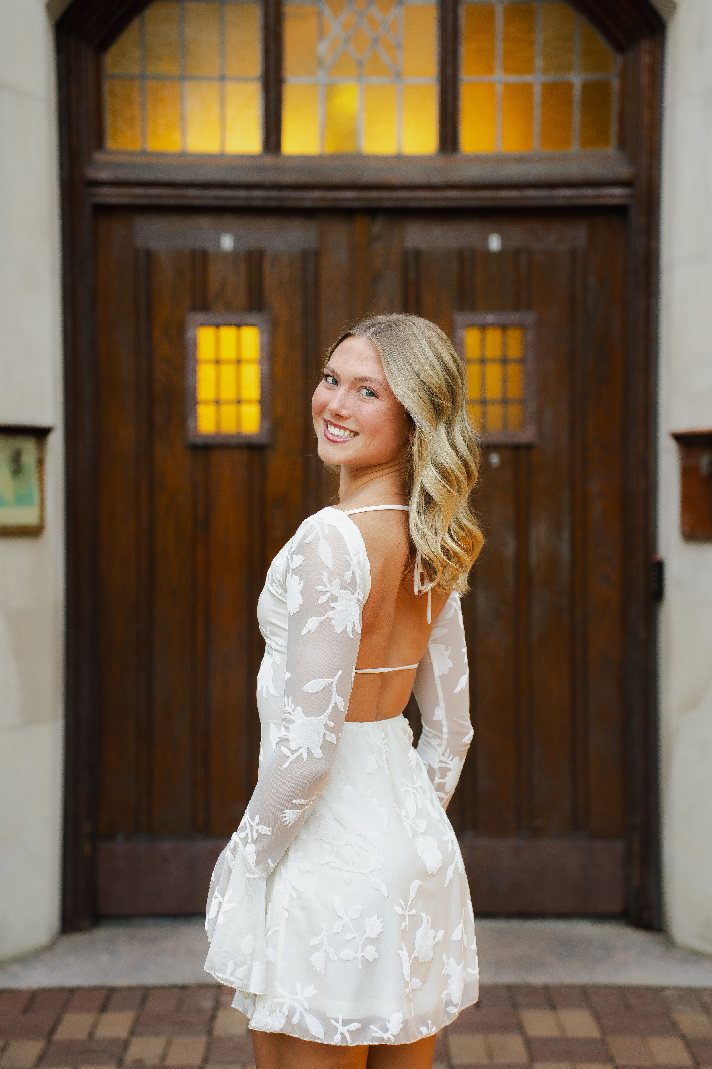 Smiling woman with wavy blonde hair looks over her shoulder in a white floral lace open-back dress in front of a rustic wooden door.