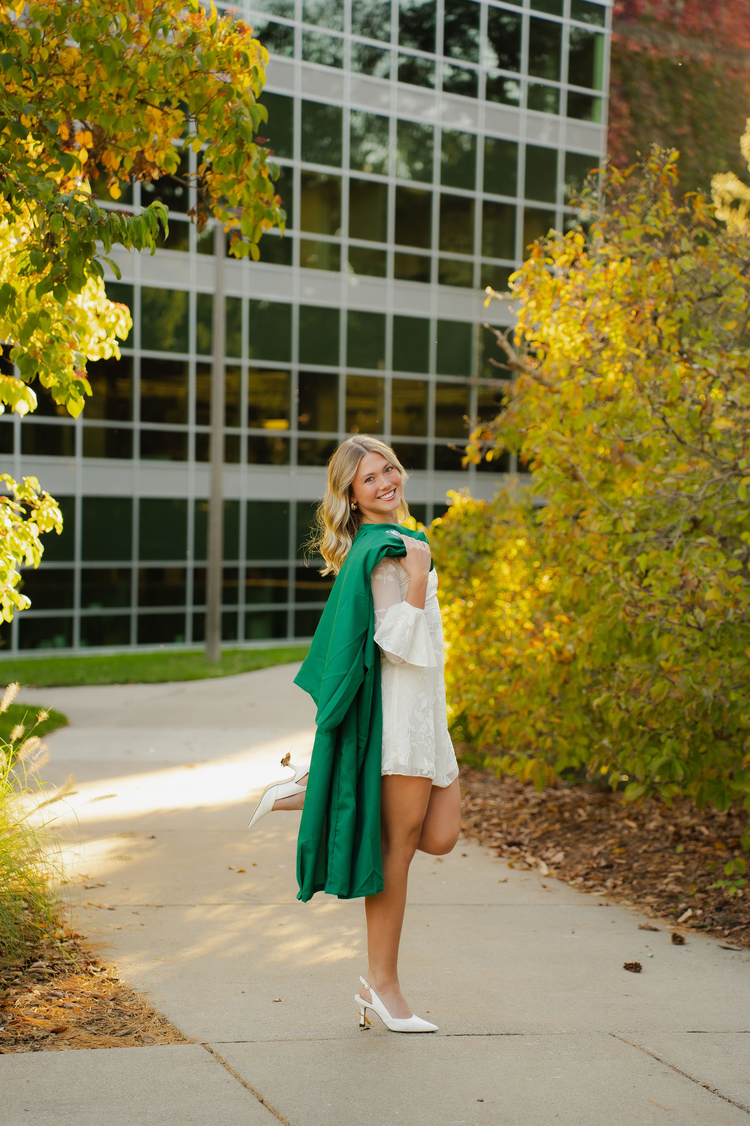 Smiling young woman in a white dress and white heels holds a green graduation gown over her shoulder on a campus path near autumn trees.