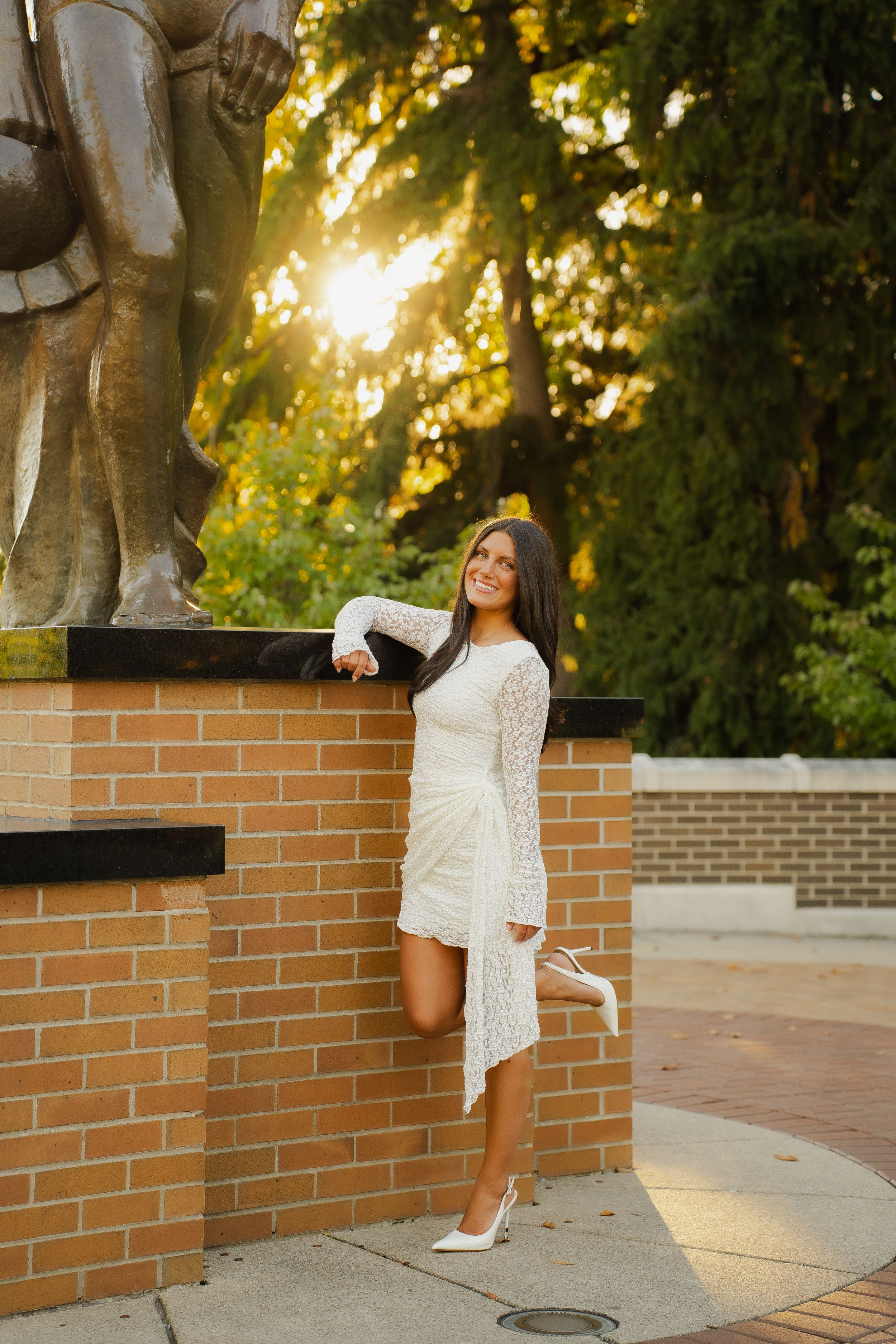 Woman in a white lace dress and white heels poses against a brick pedestal beside a bronze statue at golden hour.