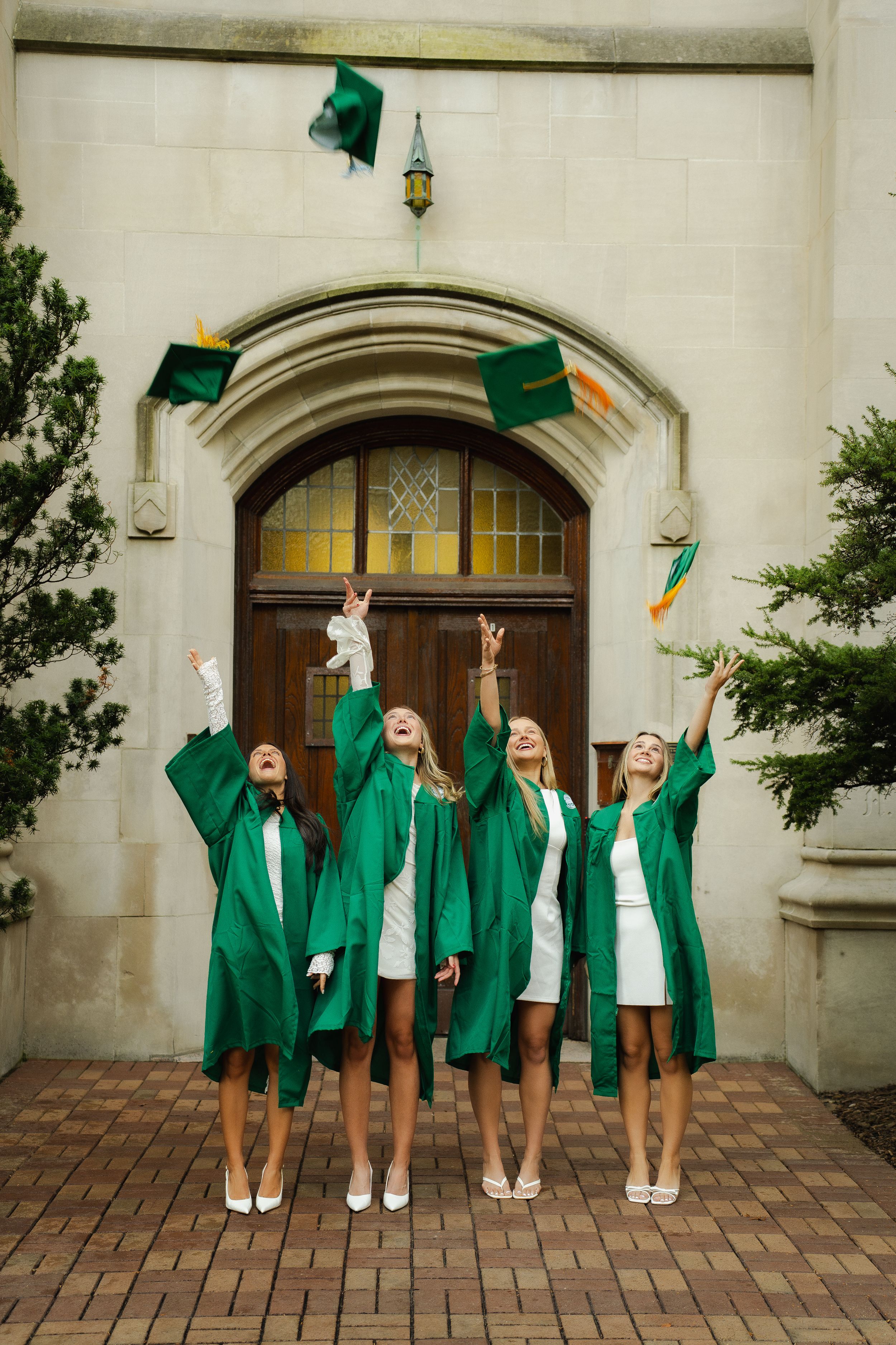 Four graduates in green gowns and white dresses toss their caps in the air in front of a historic stone building with an arched wooden door.