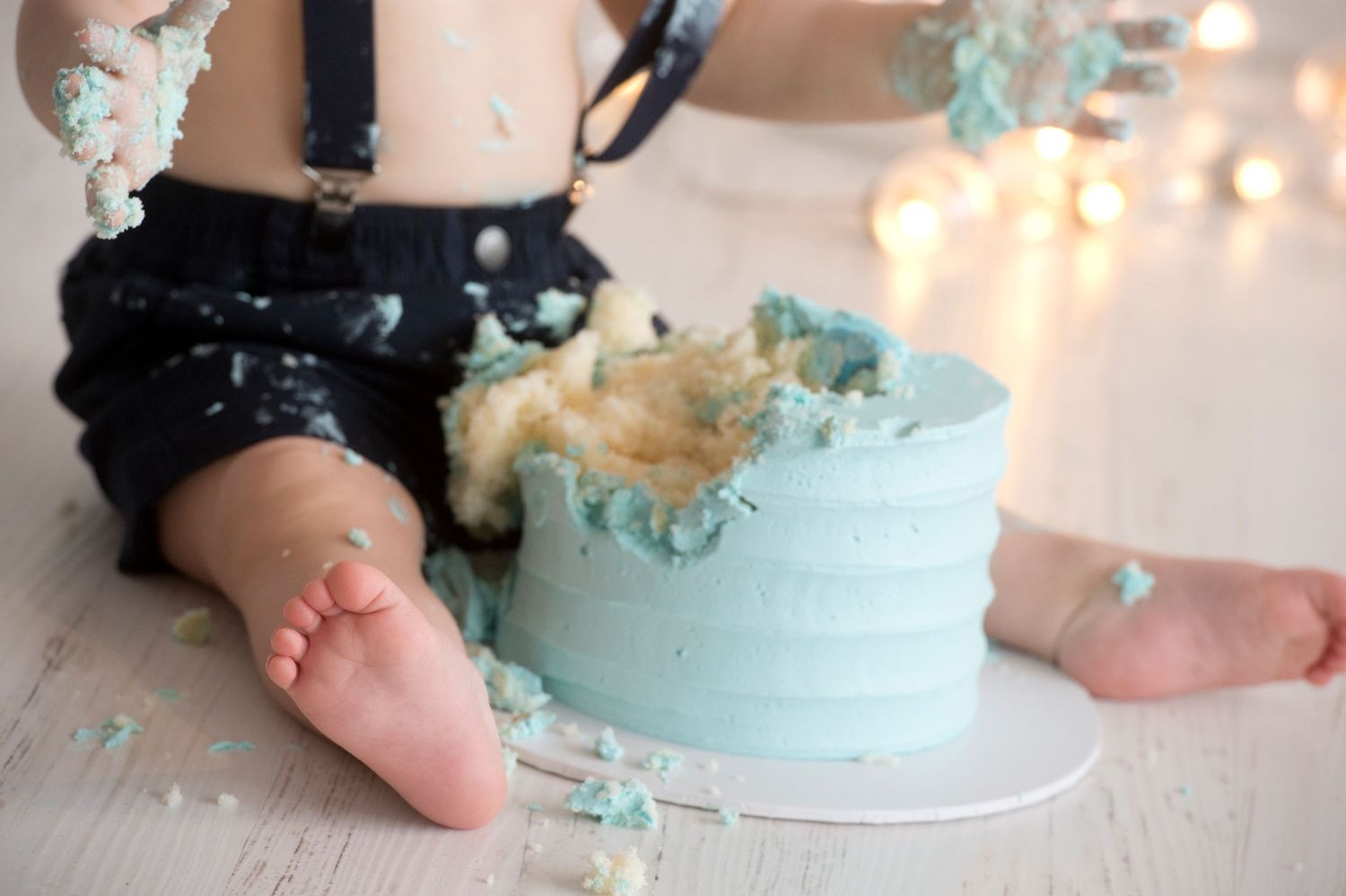 Baby's first birthday cake smash with blue frosted cake, bokeh lights background, infant in suspenders.