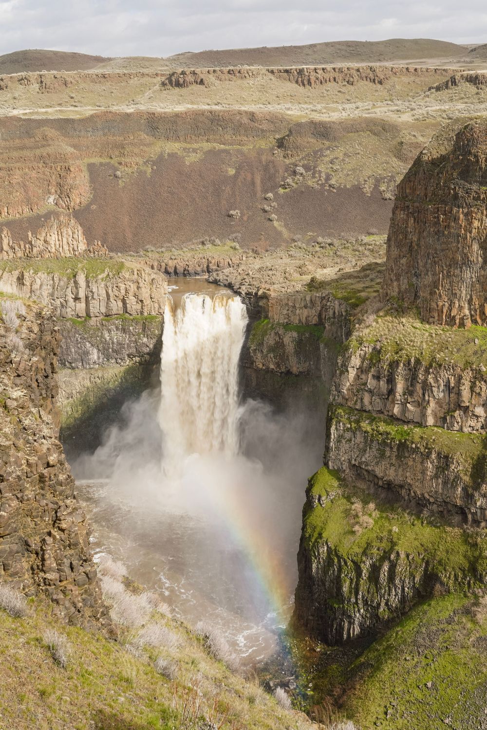A stunning waterfall cascades into a rocky canyon with a vibrant rainbow forming in the mist below.