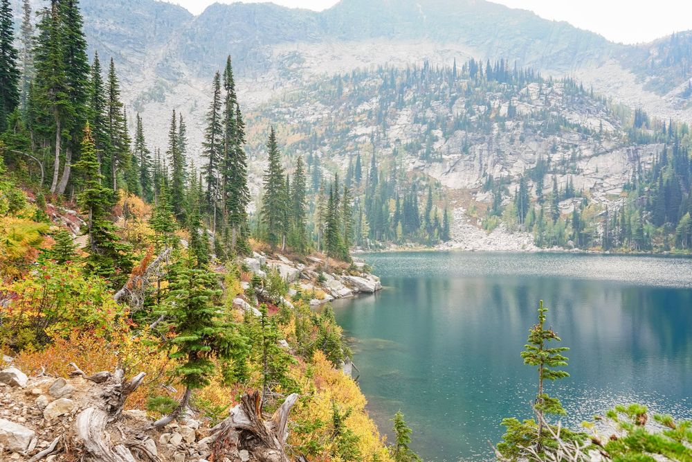 Alpine lake surrounded by evergreen trees and autumn foliage with rocky mountain slopes reflected in calm blue water.