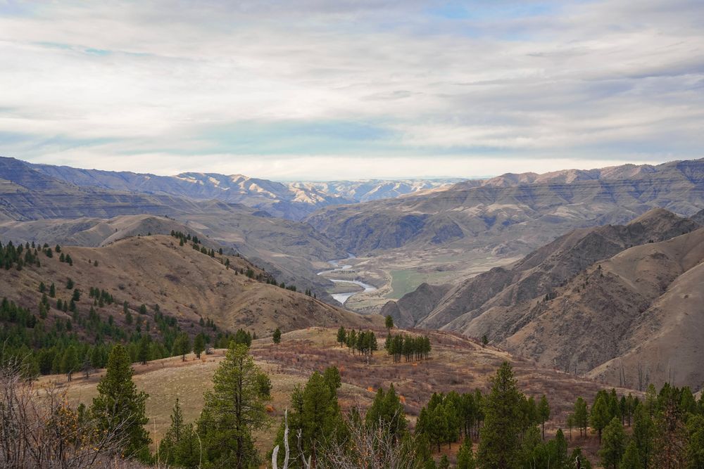 Panoramic view of a river valley surrounded by rolling hills and snow-capped mountains under a cloudy sky.