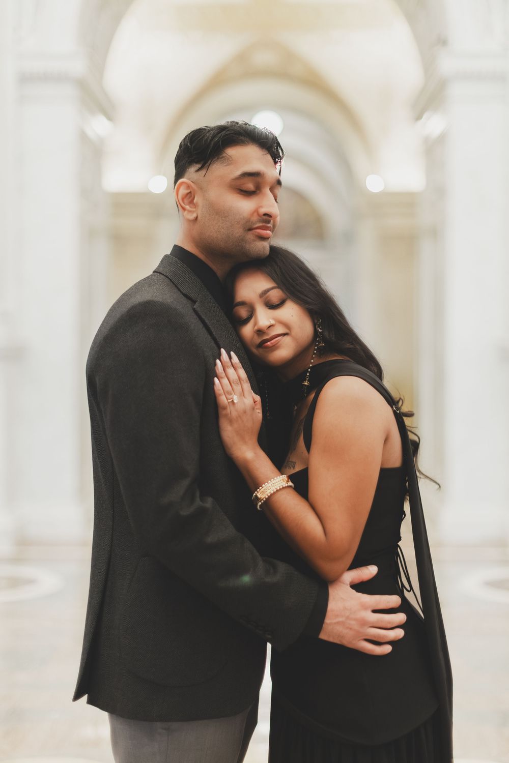 A couple in black attire share a tender embrace in an elegant white archway, eyes closed in a romantic pose.