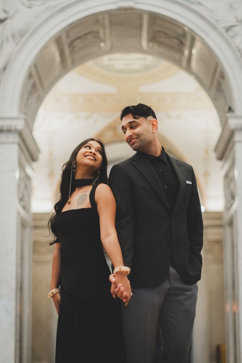 A couple in matching black outfits holds hands under an elegant marble arch in a grand classical building captured by dc engagement photo