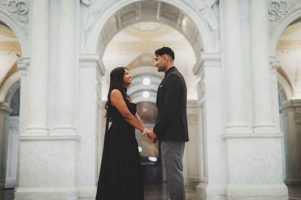 Couple holding hands facing each other in elegant white marble hall with arched columns, woman in black dress, man in gray suit.
