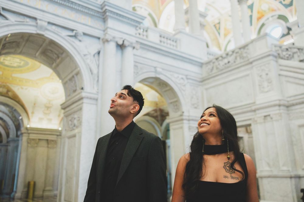 Couple in elegant black attire gazing upward inside a grand marble building with ornate arched ceilings.