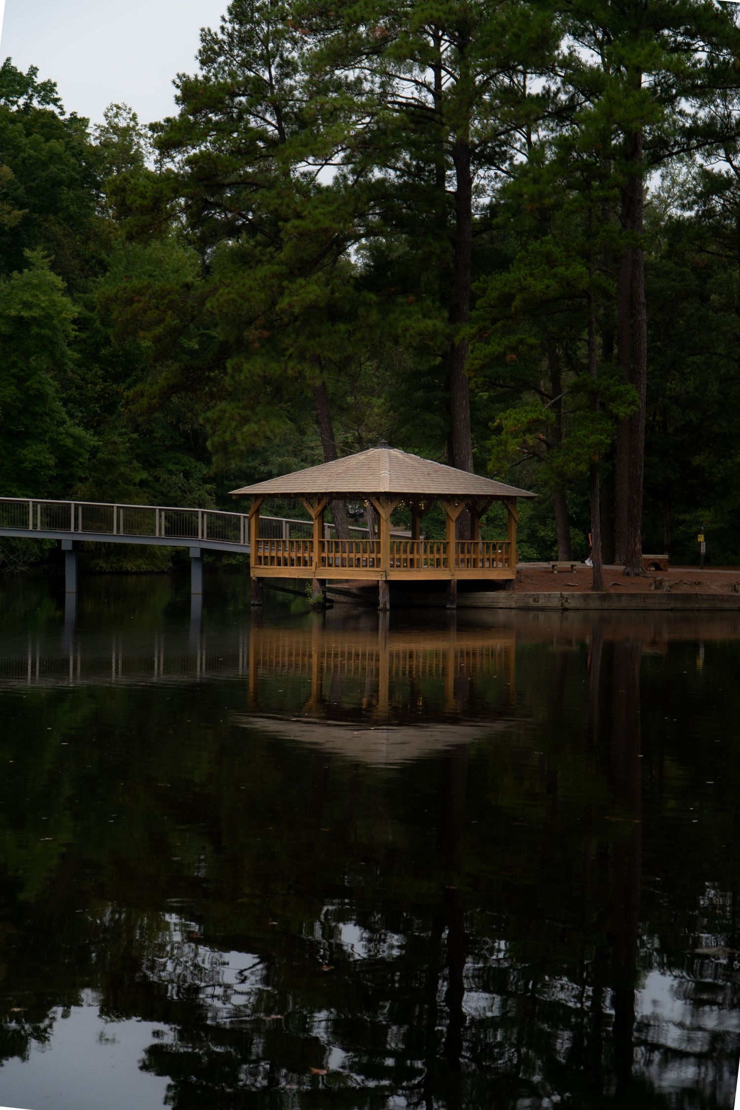 Wooden gazebo and dock structure reflected on calm lake waters surrounded by tall pine trees.