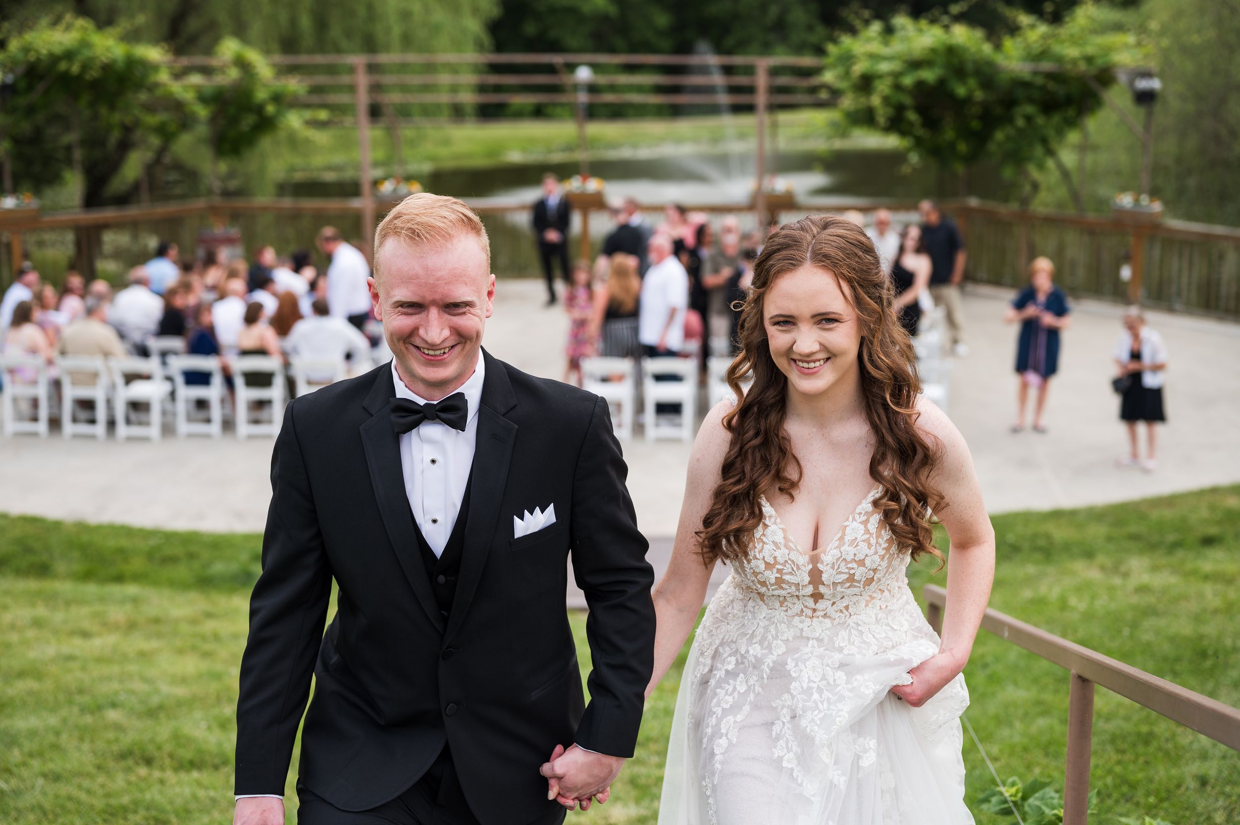 Bride and groom holding hands walking away from outdoor wedding ceremony on lush green lawn.
