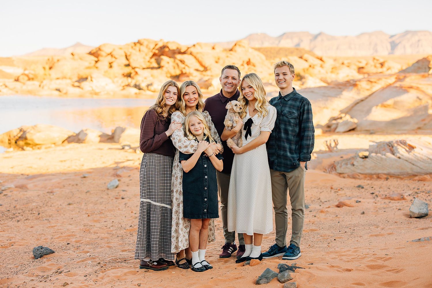 Family of six smiling together on red desert terrain with scenic lake and rock formations behind them.