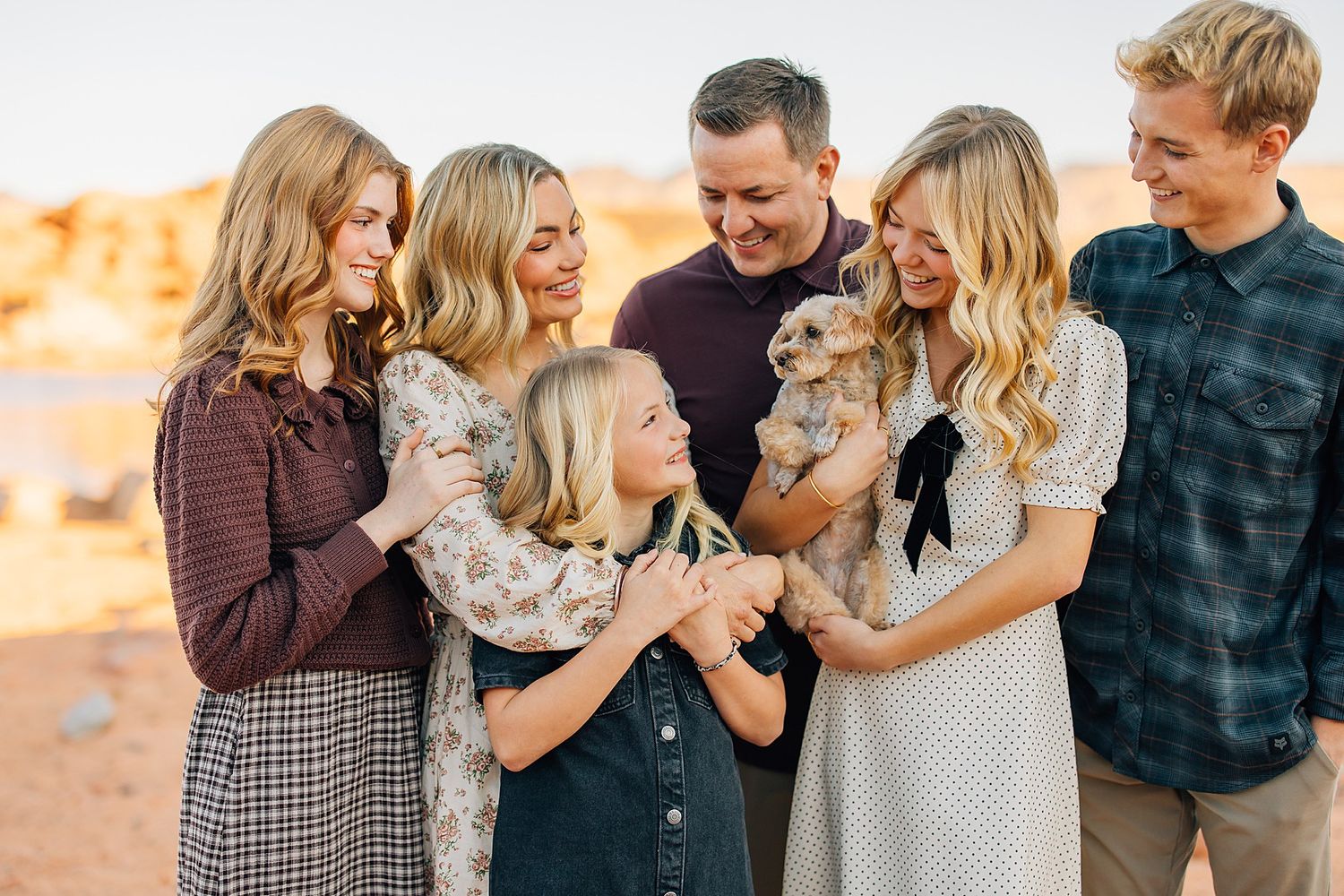 A joyful family of six gathers outdoors at sunset, smiling and holding a small dog in a warm desert setting.