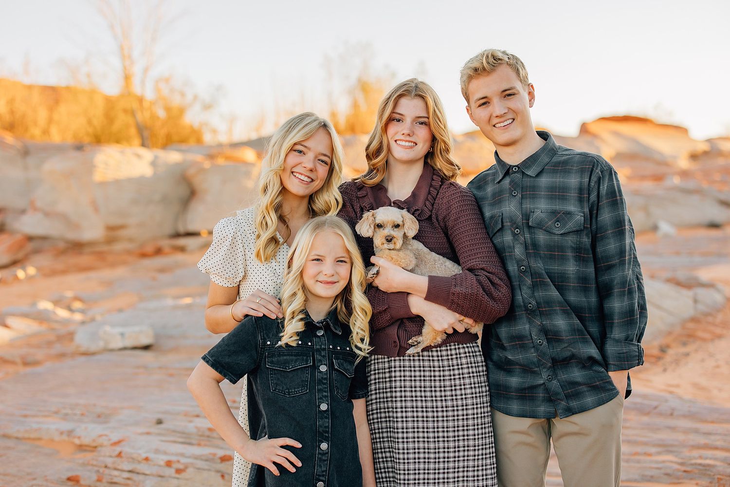 Three siblings and a young girl holding a small dog pose outdoors near red rock formations at sunset.