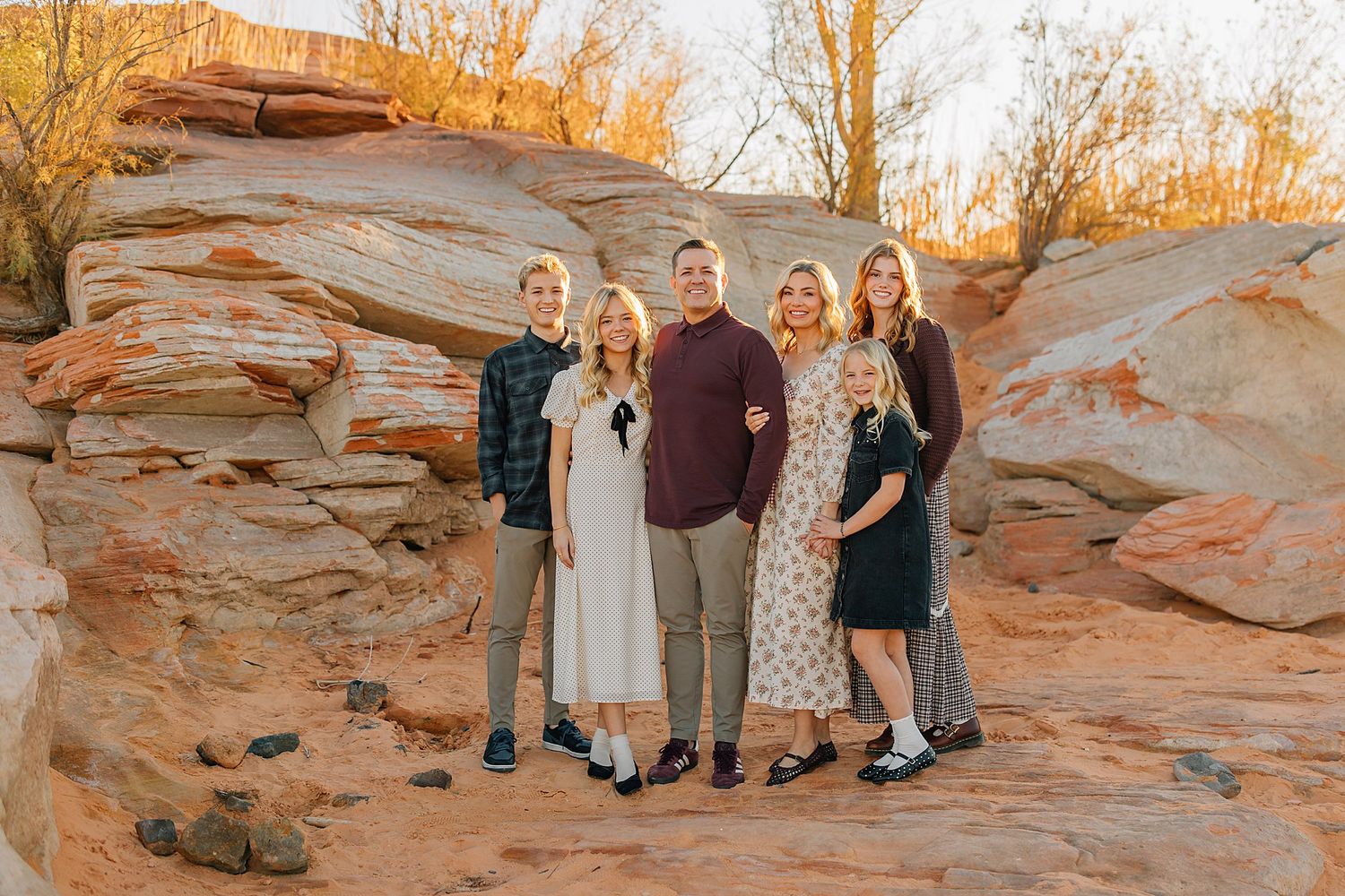 Family of five smiling together among red rock formations at golden hour in a desert landscape.