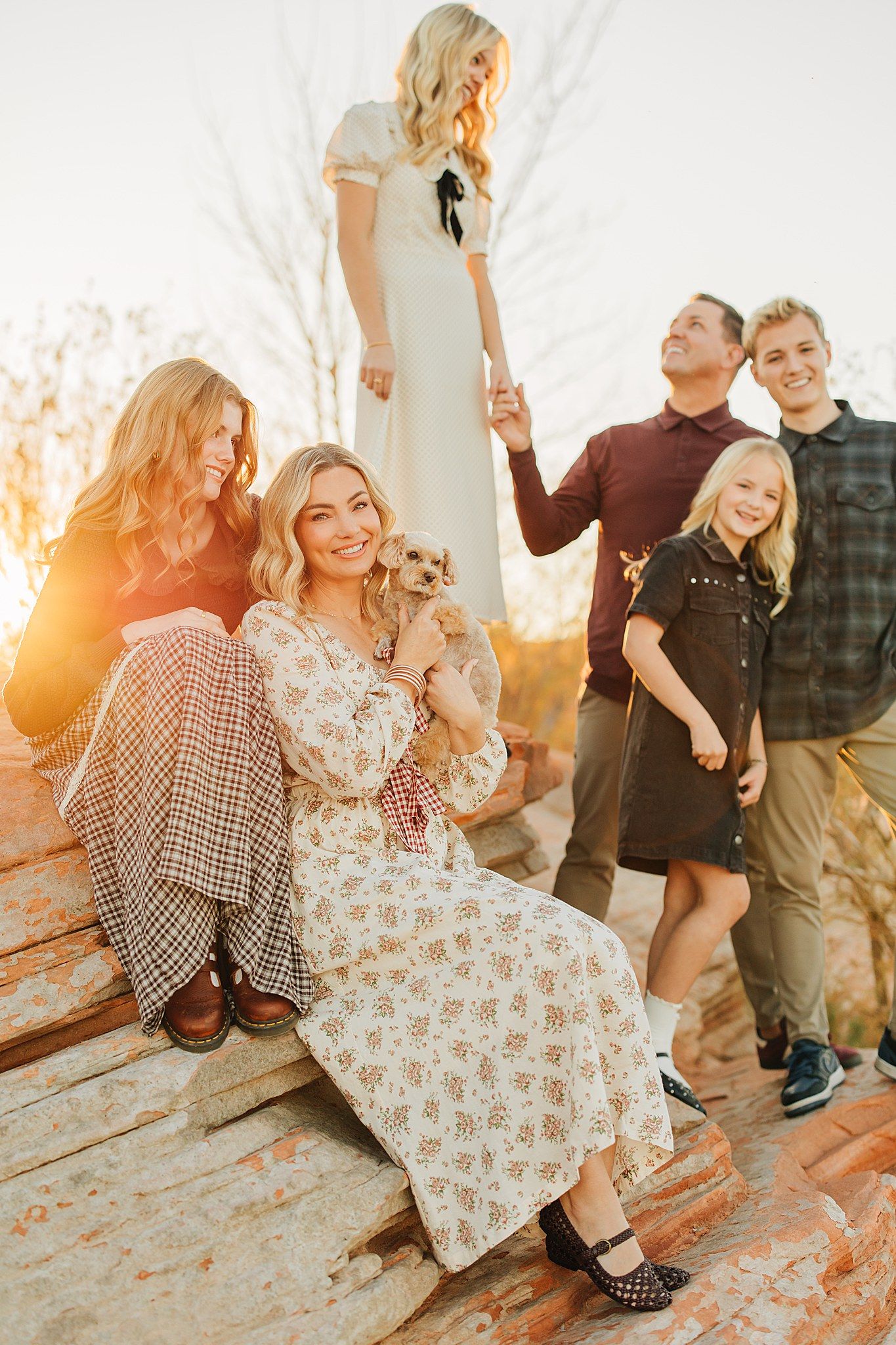 Family of six with small dog posing on red rock formations during warm golden sunset outdoors.