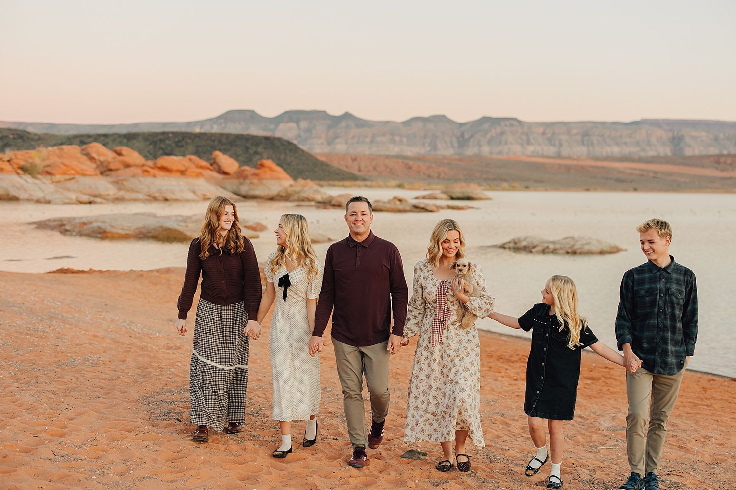 Family of six walking hand-in-hand on red desert landscape with lake and mountains at sunset.