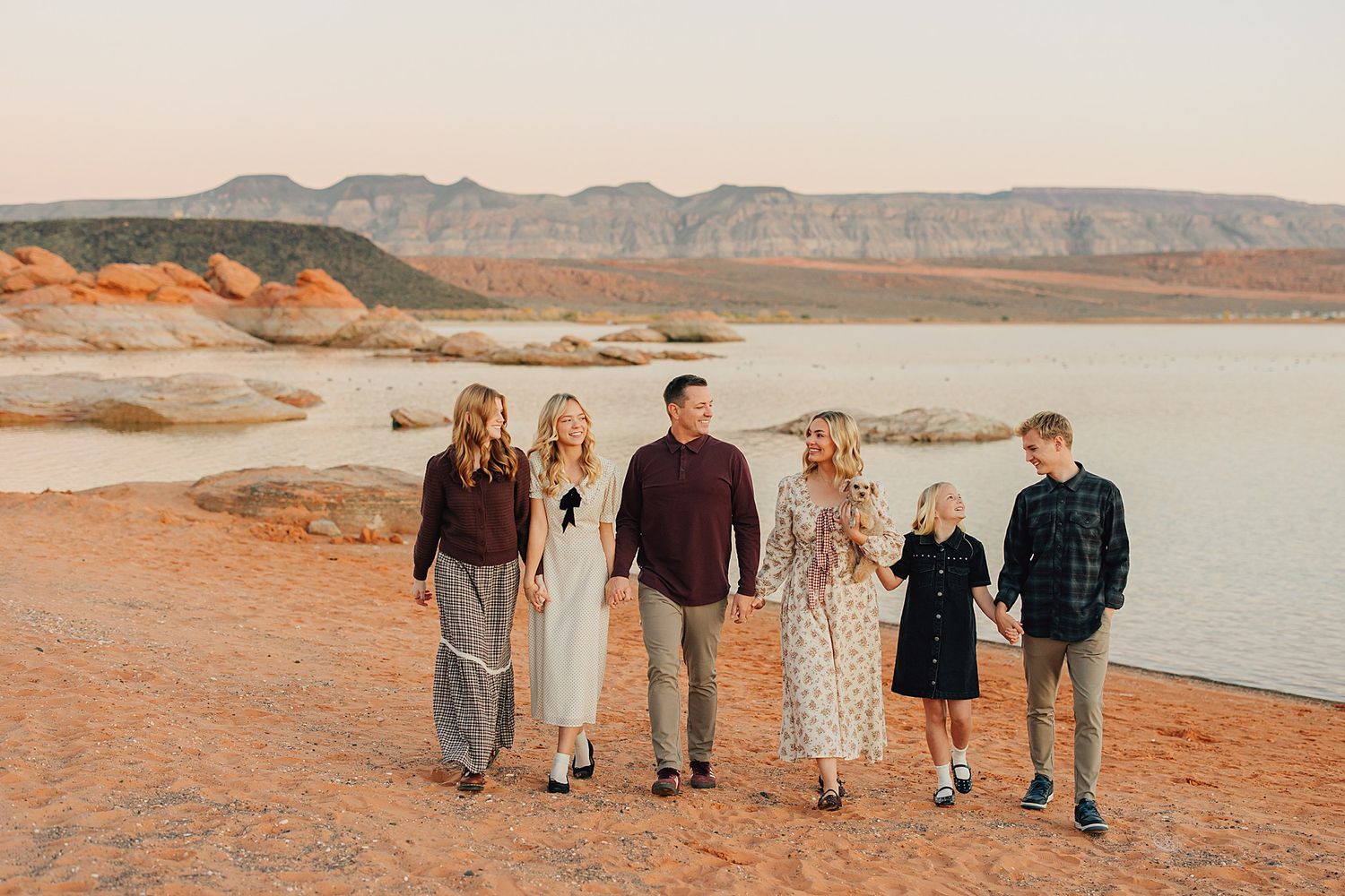 Family of six walking along a red sand shoreline with mountains and calm water in the background at sunset.