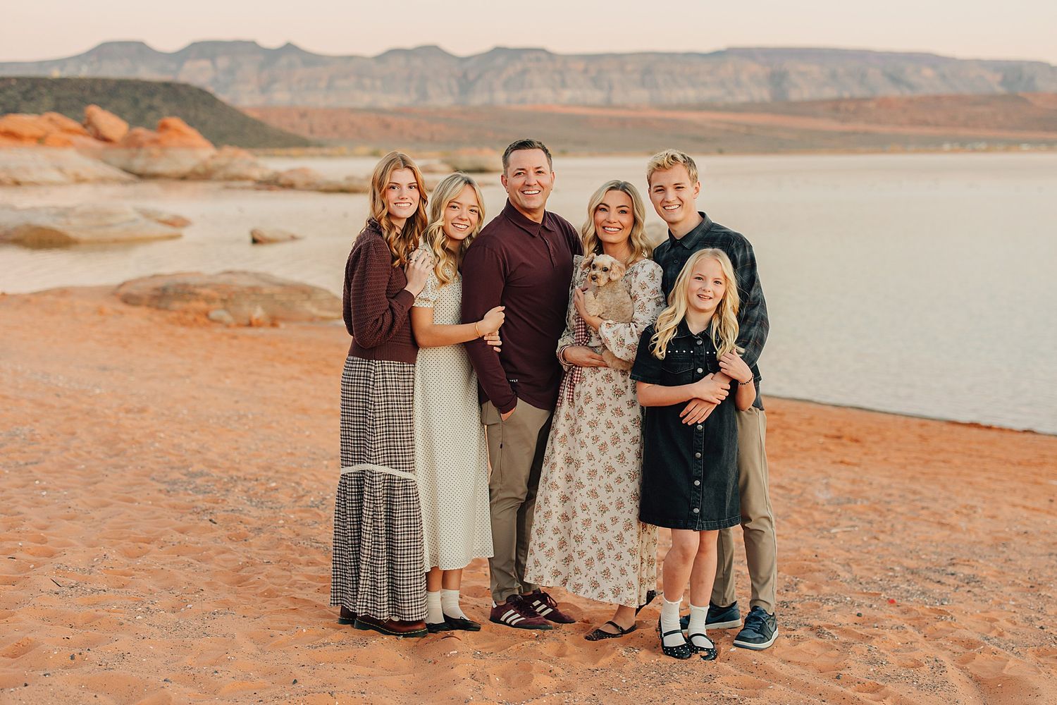 Family of six smiling together outdoors near a lake with red rock formations and mountains in the background.