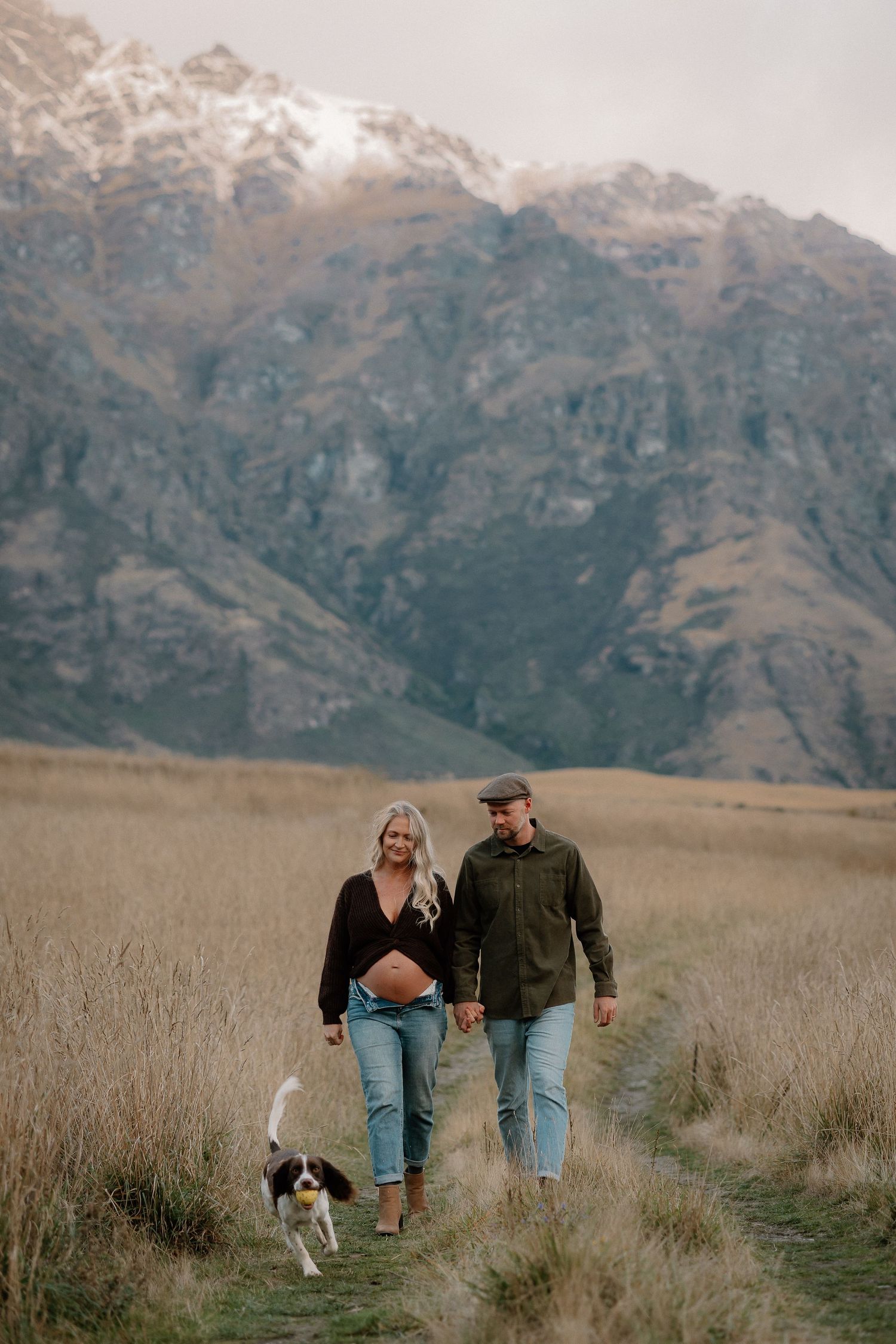 Pregnant couple walking with their dog through a mountain field in New Zealand, holding hands against snowy peaks.