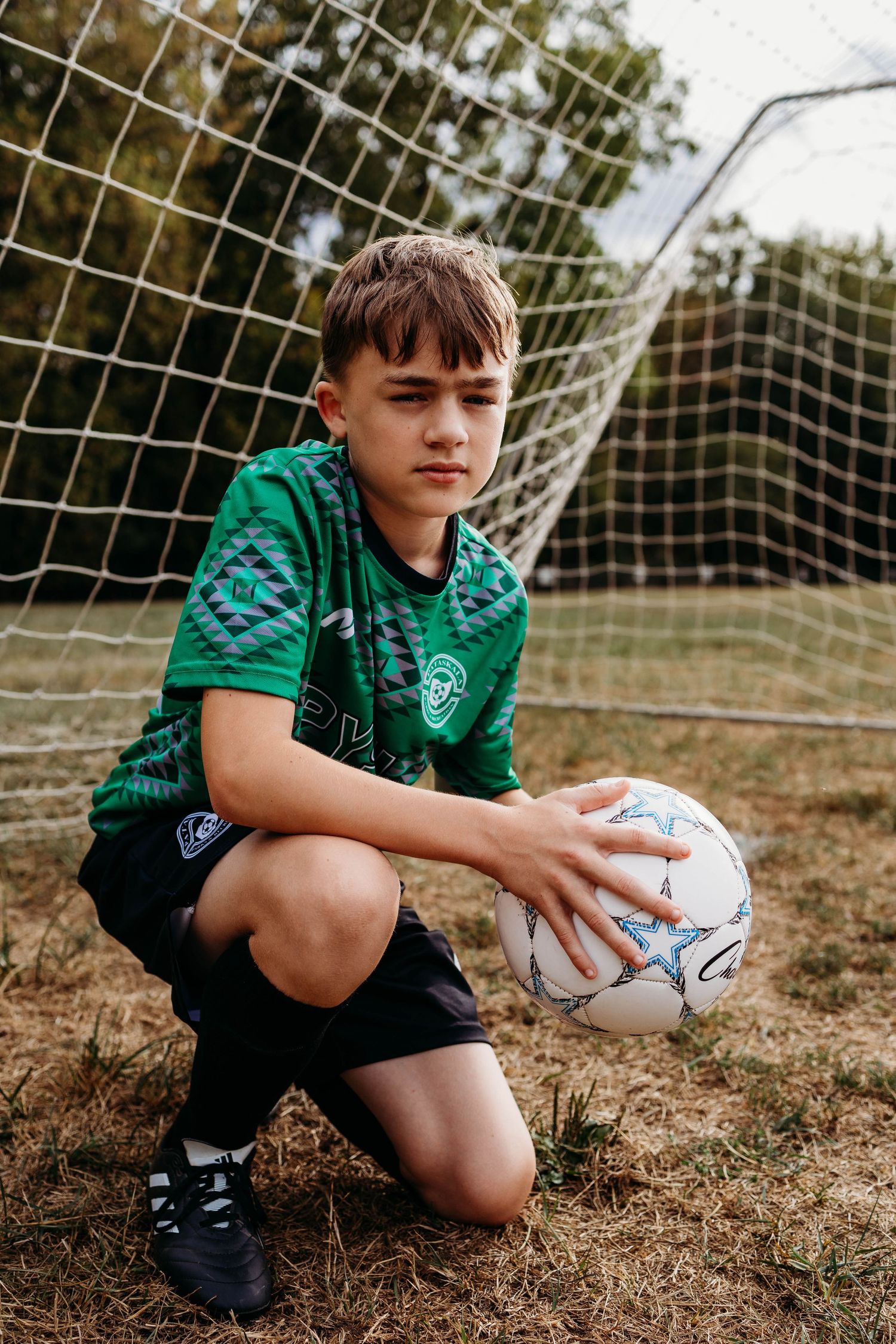 Young soccer player in green jersey kneels with ball in front of goal net during practice session.