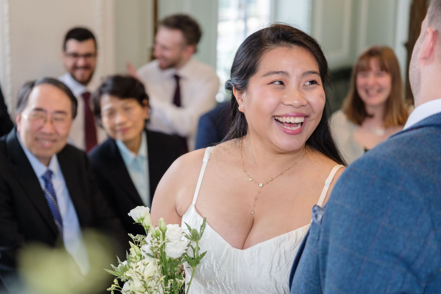 Smiling bride holding white flowers during wedding ceremony with guests in background.